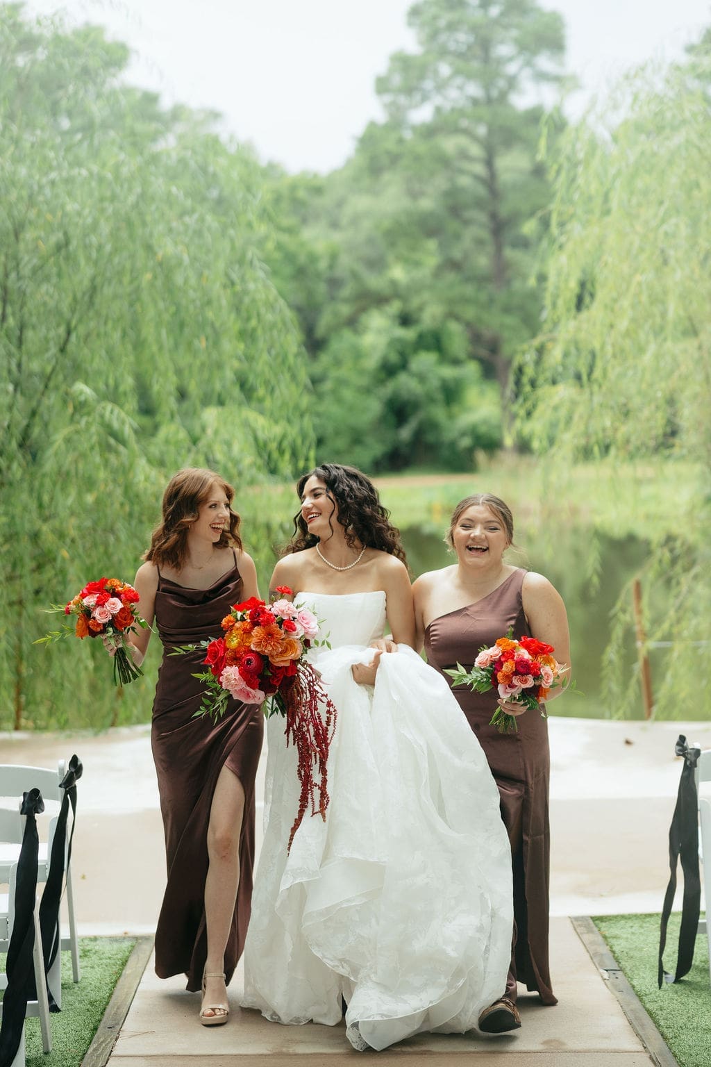 Bride walks down the aisle outdoors with her bridesmaids in brown dresses, all holding vibrant red and orange bouquets and laughing together.