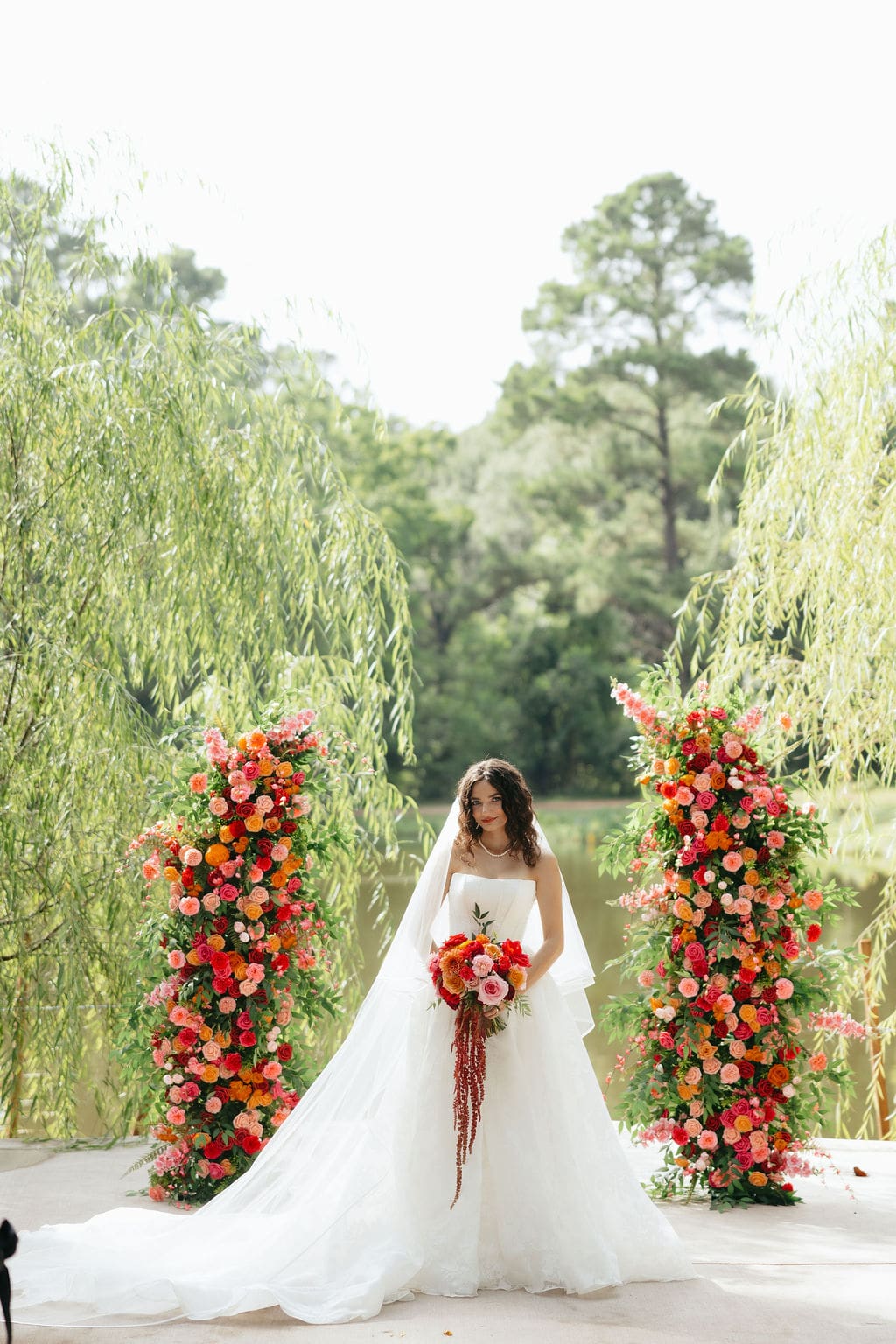 Bride stands between two tall floral arrangements filled with red, orange, and pink blooms beside a lake, a striking portrait blended seamlessly into documentary wedding photos coverage.