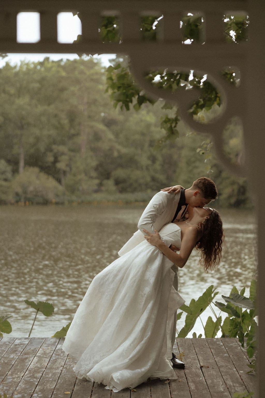 A candid wedding photo of a groom dipping his wife for a kiss while they stand by a pavilion by a pond while it was pouring rain during the wedding portraits.