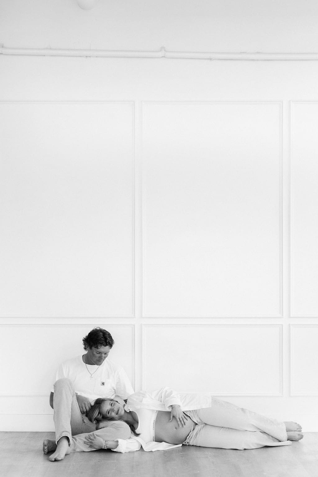 Soon to be parents sitting on the floor during the studio maternity photos, with the mom laying down with her head on her husbands lap as she looks at the camera and holds her belly.