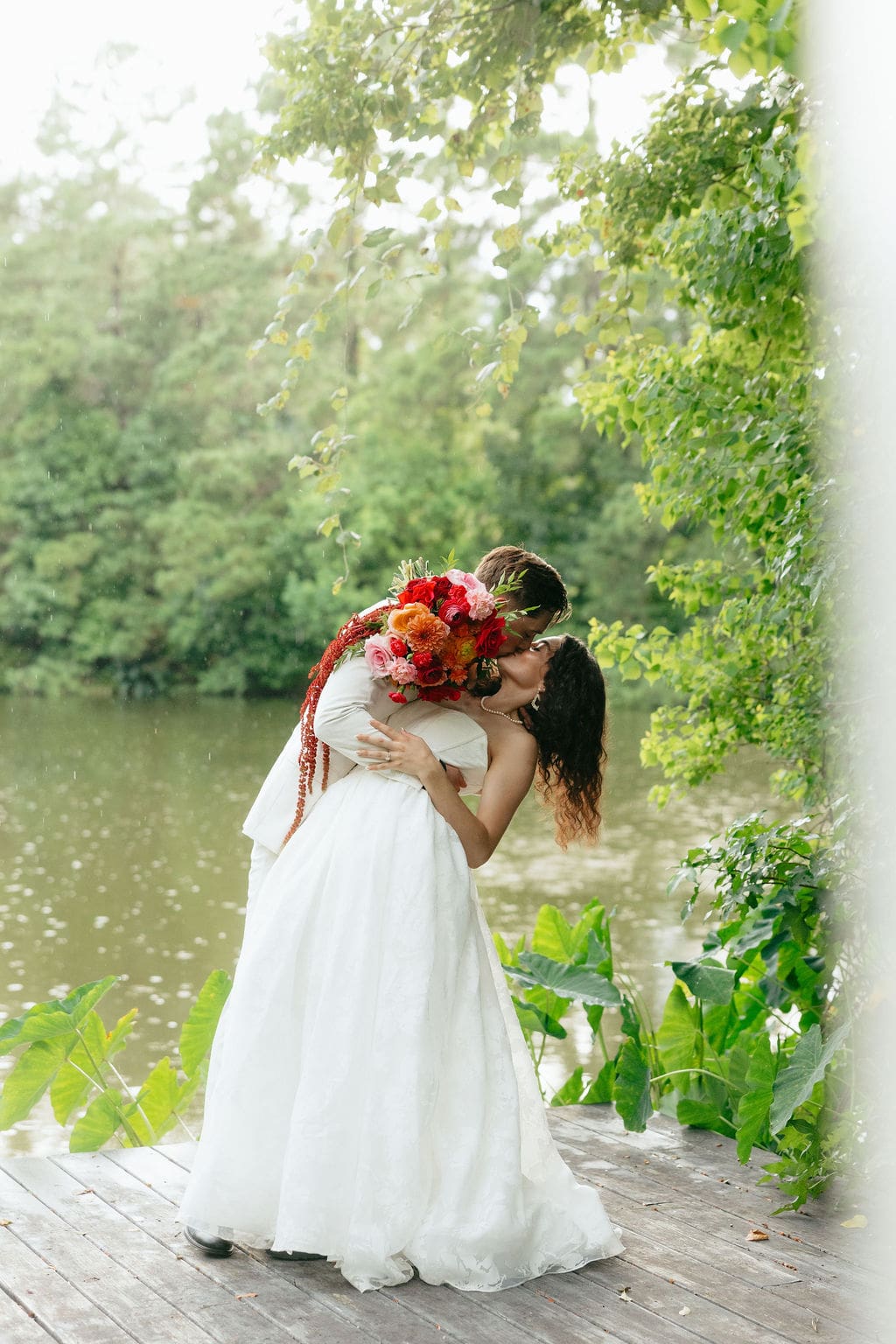 A candid wedding photo of a groom dipping his wife for a kiss while they stand by a pavilion by a pond while it was pouring rain during the wedding portraits.