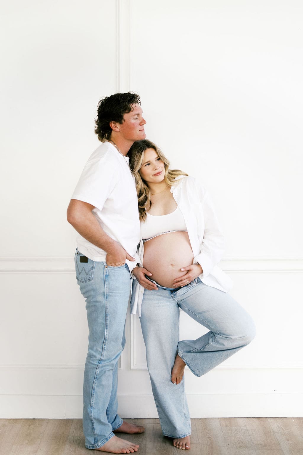A studio couple photo of the soon to be parents standing while the mom holds her belly while they look away from the camera.