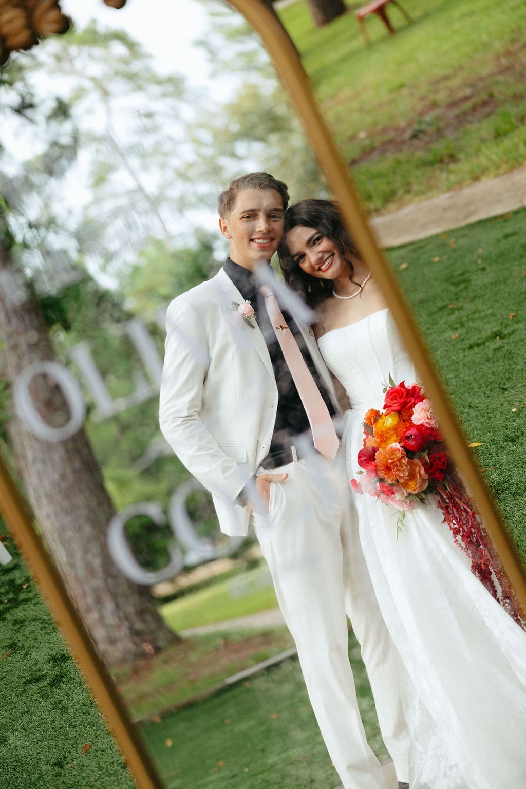 Bride and groom smile together in a mirror reflection outdoors, holding a bold bouquet of red and orange flowers on their wedding day.
