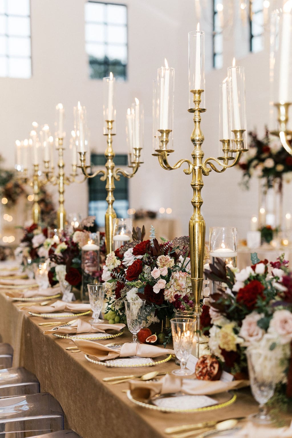 A close-up of tall gold candelabras and lush burgundy and blush floral centerpieces lining the reception dinner table at Boxwood Manor.
