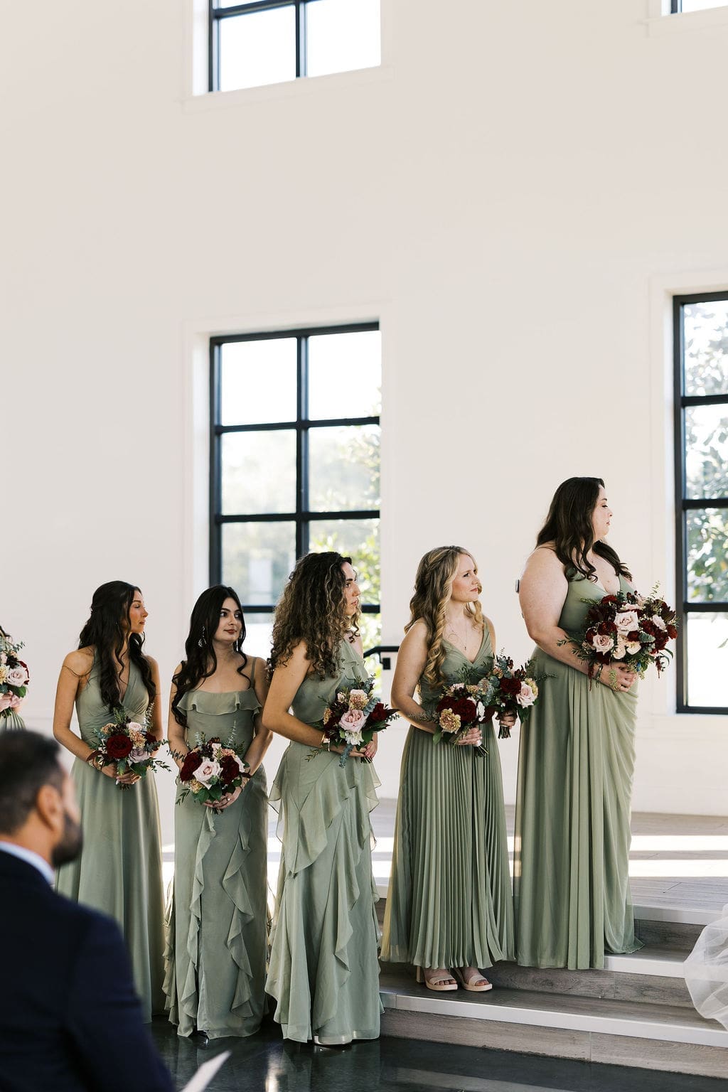Five bridesmaids in sage green gowns holding deep red and blush bouquets stand at the altar during the ceremony at Boxwood Manor Houston.