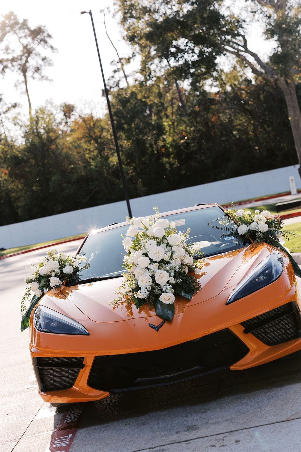 An orange Corvette decorated with a lush white rose and greenery floral arrangement on the hood serving as the getaway car at Boxwood Manor Houston.