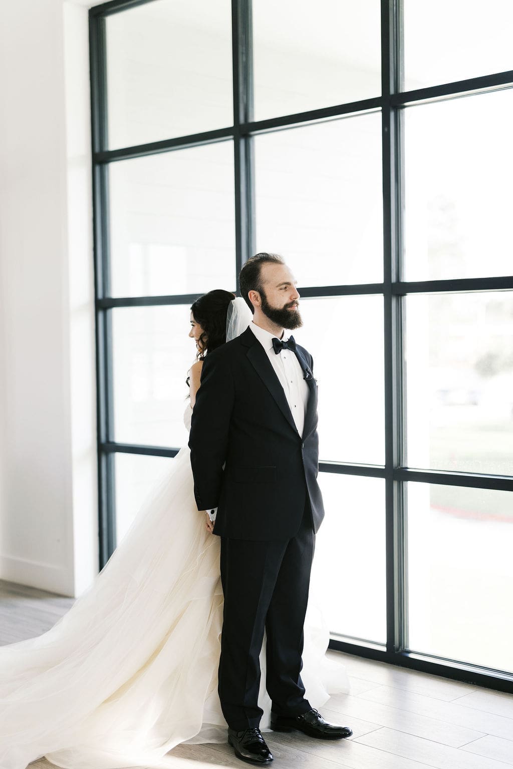 The bride and groom stand back to back in front of a dramatic floor-to-ceiling black-framed window at their Persian wedding at Boxwood Manor.