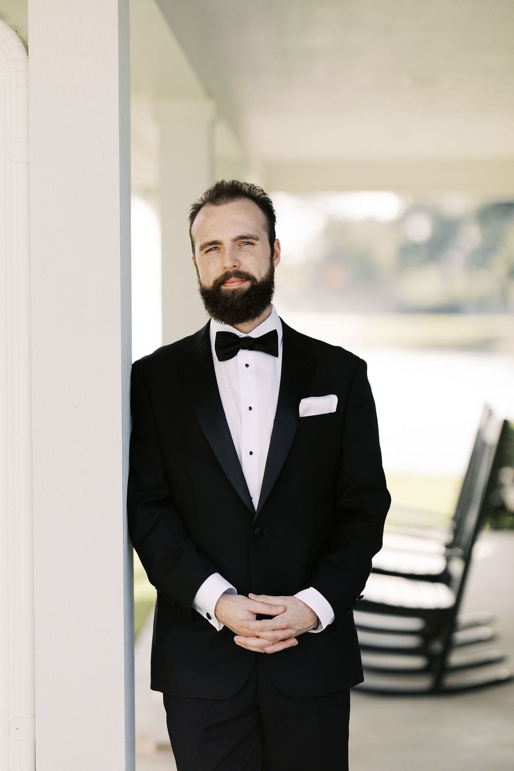 The groom in a classic black tuxedo and bow tie leans against a white column on the porch at this Persian wedding venue in Houston.