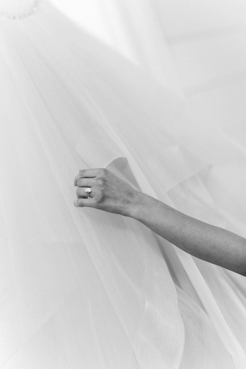 A black and white close-up of the bride's hand wearing a diamond engagement ring while holding her flowing wedding veil.