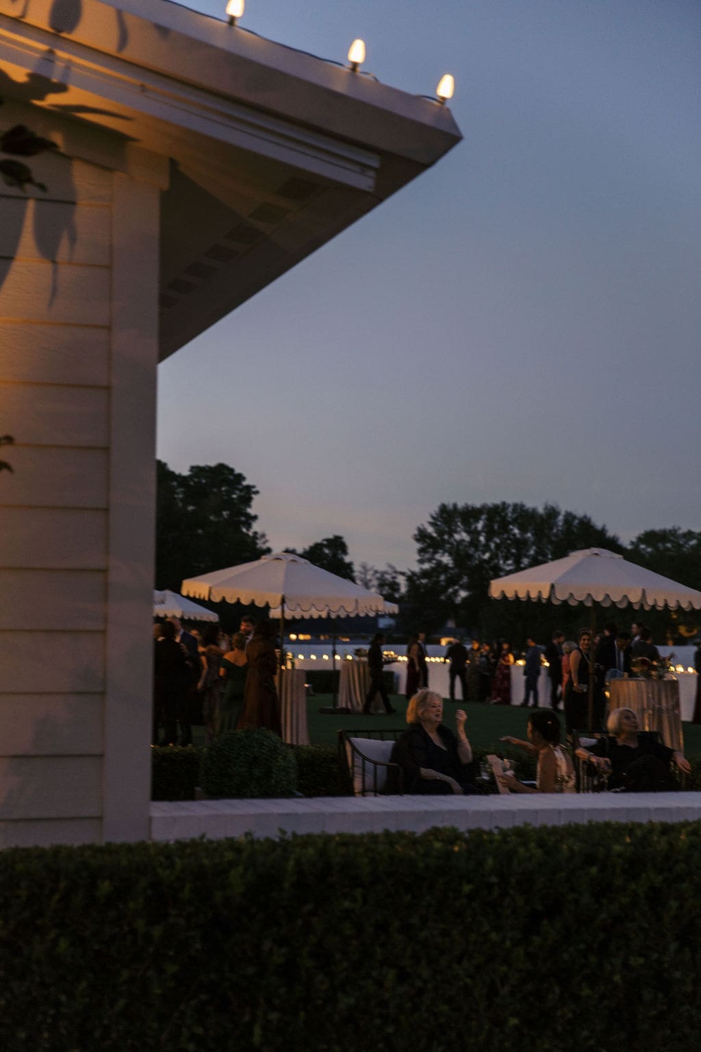 Wedding cocktail hour guests mingle on the lawn under white market umbrellas at dusk during the Persian wedding at Boxwood Manor Houston.
