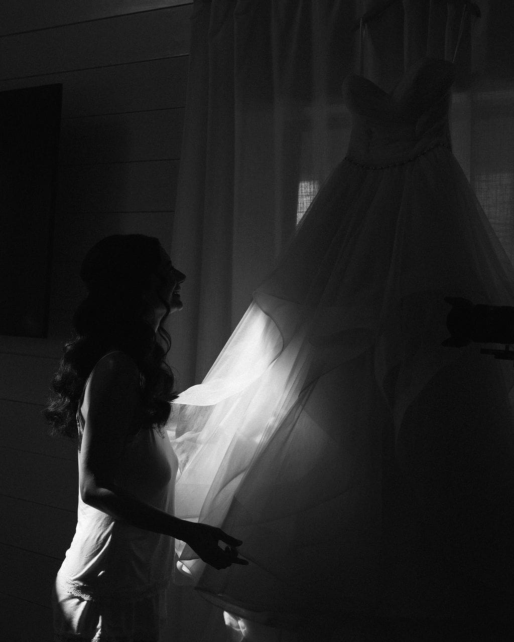 A dramatic black and white silhouette of the bride gazing at her wedding gown hanging in the window light.