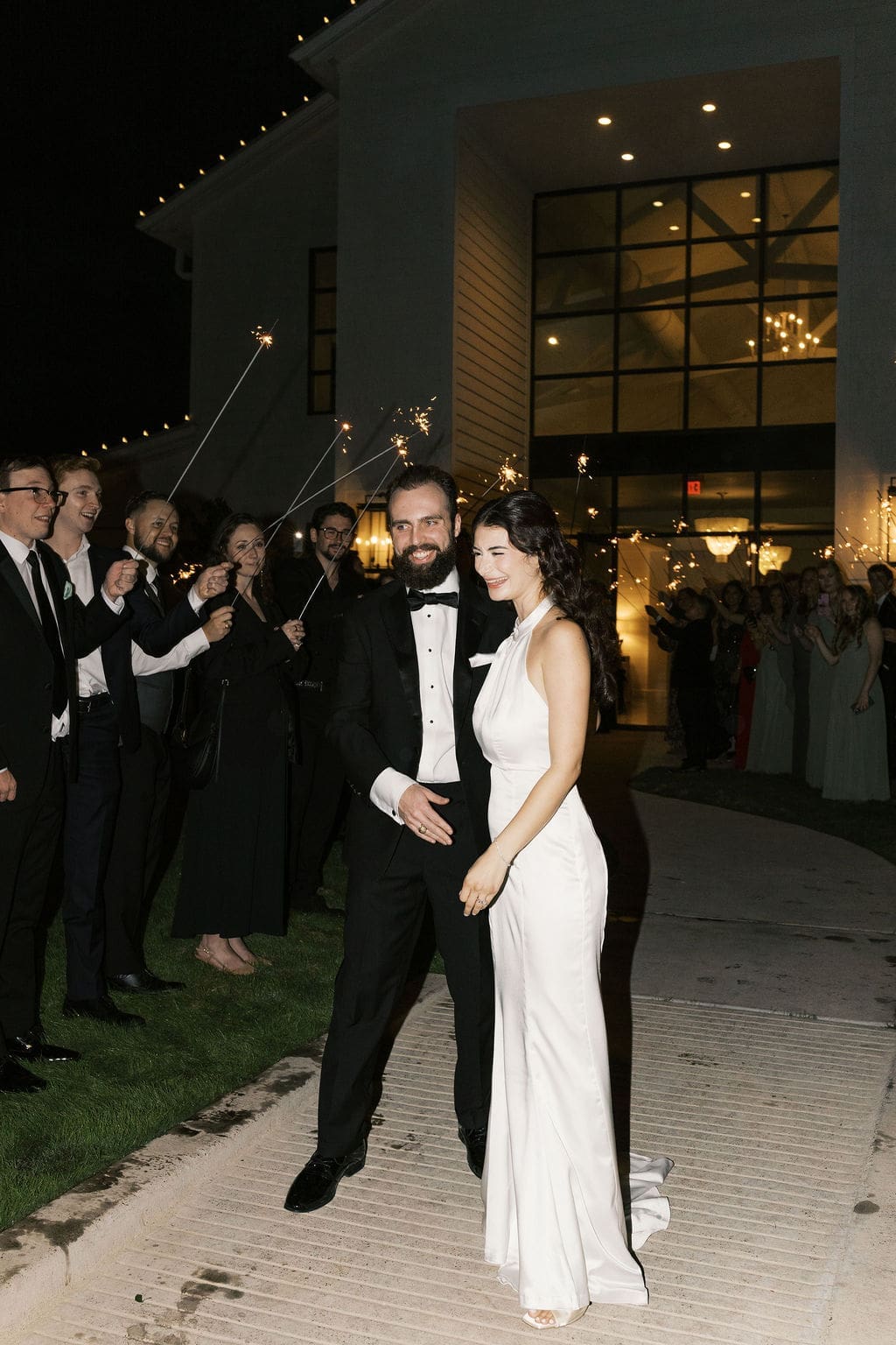 The bride and groom walk through a glittering sparkler sendoff lined with cheering guests outside the illuminated Boxwood Manor at night.