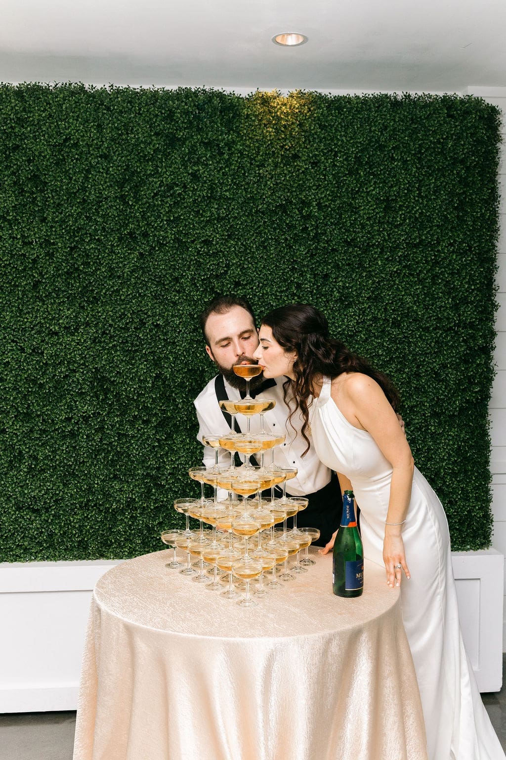 The newlyweds playfully sip from the top of their champagne tower together in front of the boxwood hedge backdrop at their Persian wedding reception.