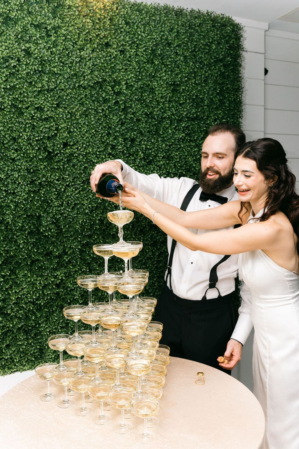 The bride and groom laugh together while pouring champagne down a coupe tower in front of a green boxwood hedge wall at the reception.