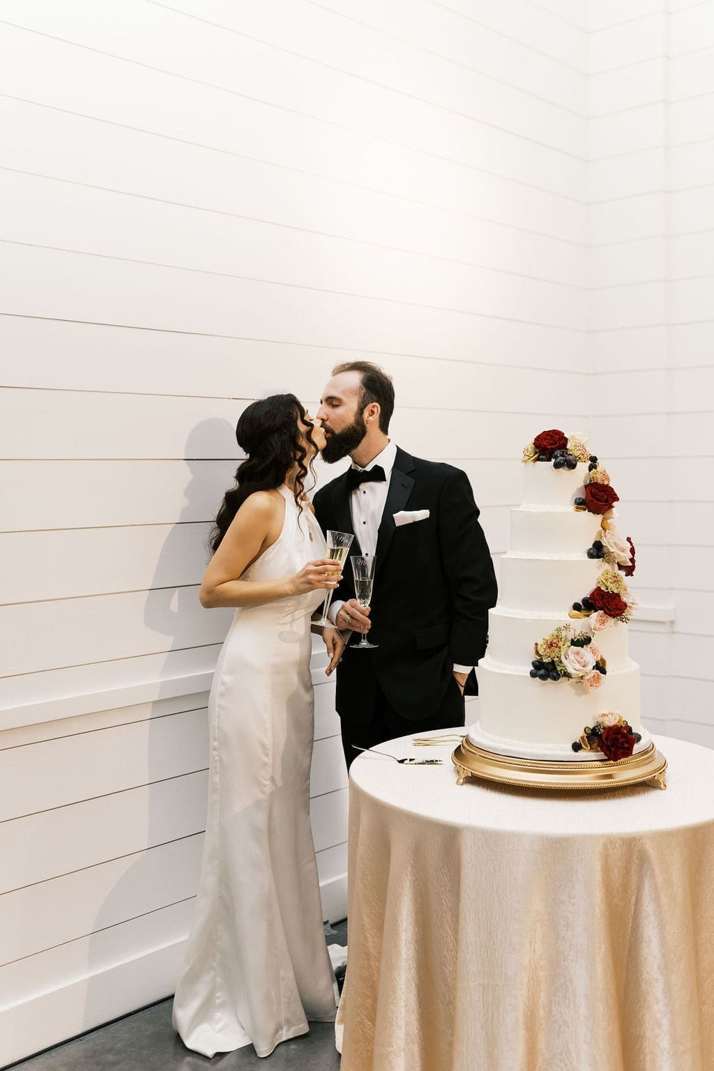 The bride and groom share a kiss beside their white five-tier wedding cake decorated with cascading red and blush flowers at their Persian wedding reception.