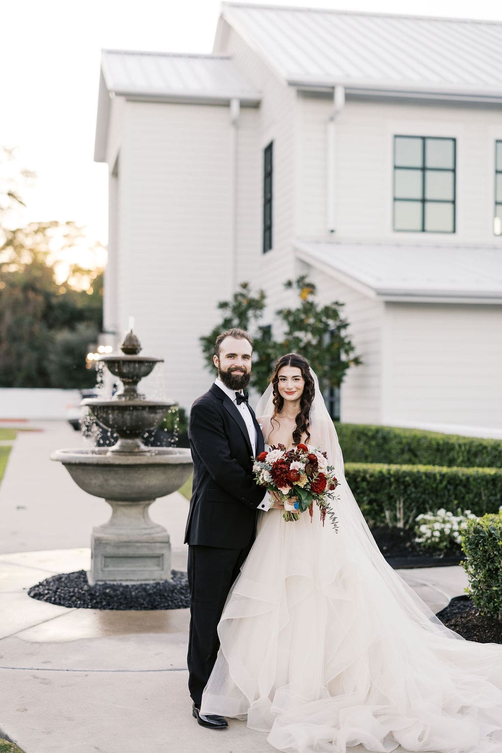 The bride and groom pose together in front of a stone fountain outside the white manor venue at golden hour.