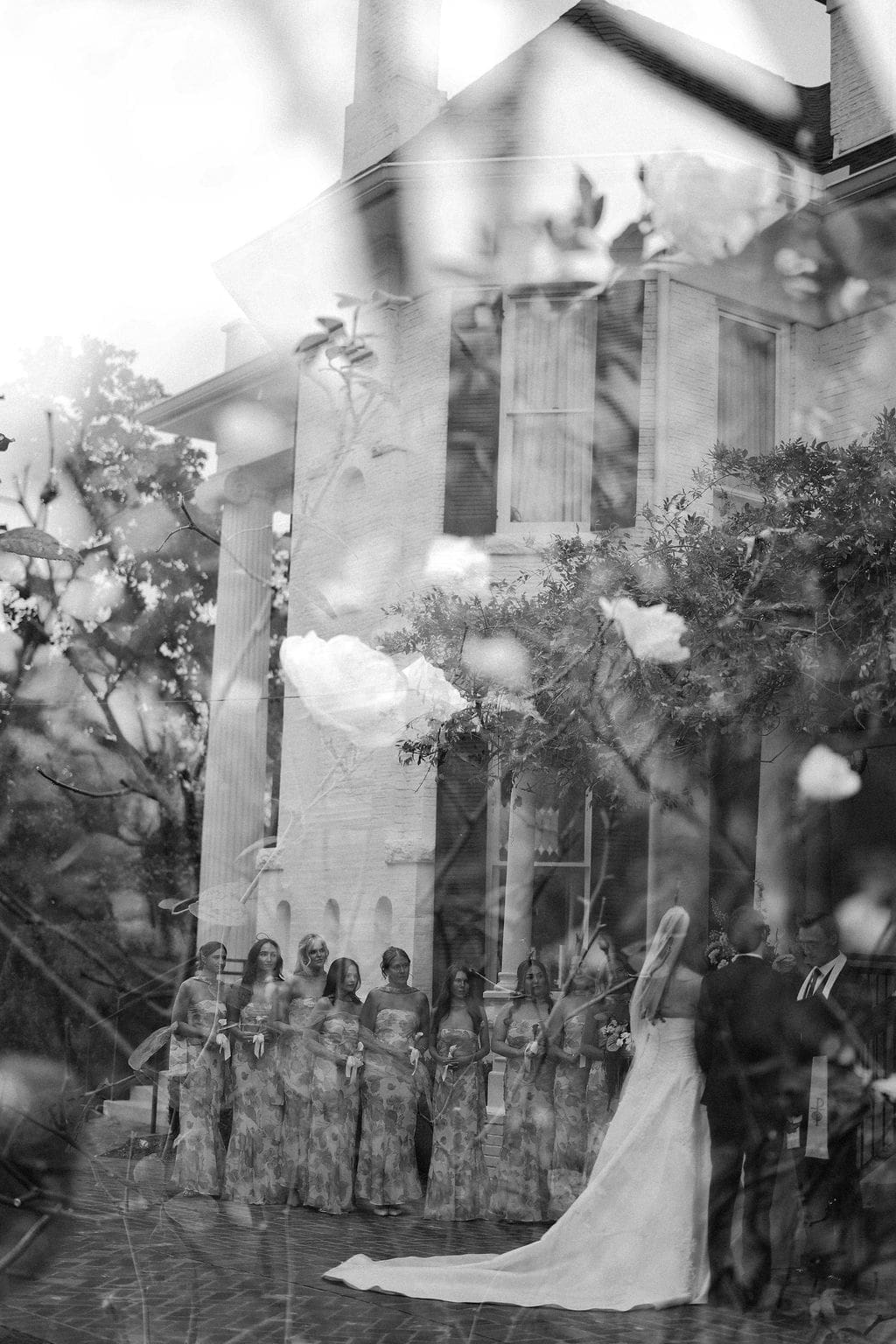 Black and white photograph of a bride and bridesmaids standing outside a historic estate during the ceremony.