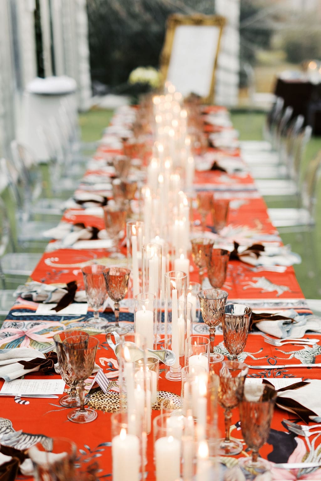 Long reception table decorated with patterned linens, crystal glassware, and tall candles arranged for an outdoor dinner.