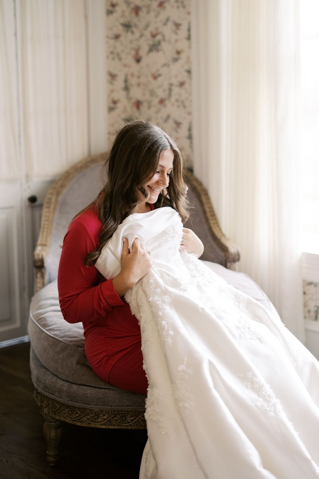 Bride in a red getting-ready outfit sitting in a vintage chair, smiling while holding her lace wedding dress in a softly lit bridal suite.
