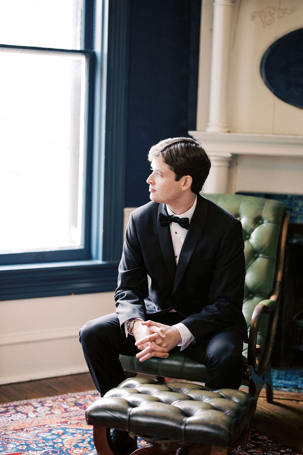Groom sitting by a window in a green leather chair, getting ready in a classic tuxedo before the wedding ceremony.