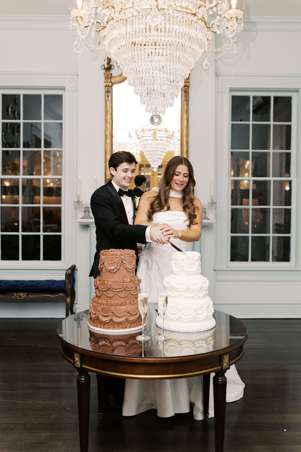 Bride and groom cutting their wedding cake beneath a crystal chandelier during an elegant Mansion Wedding celebration.