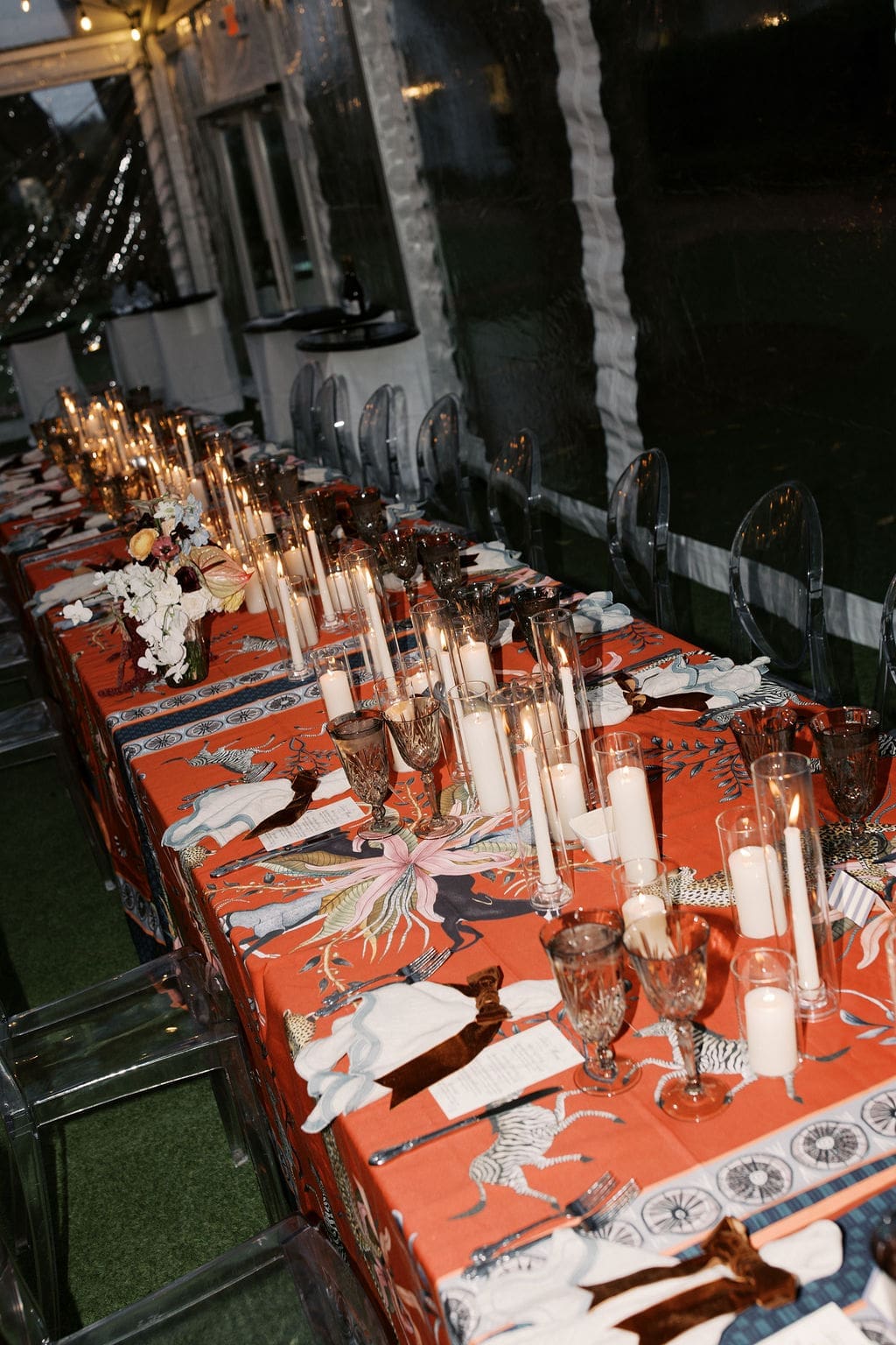 Evening reception table design with tall glass candle holders, patterned table linens, and clear chairs under a tent.