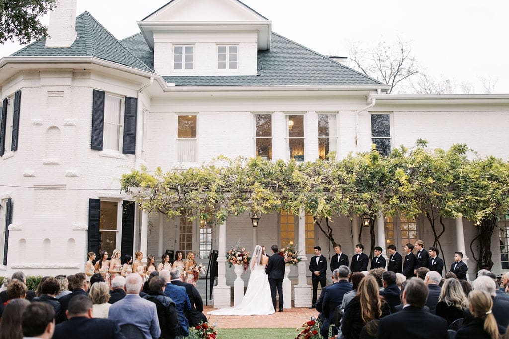 Outdoor wedding ceremony taking place in front of a grand white estate with guests seated and the couple exchanging vows.