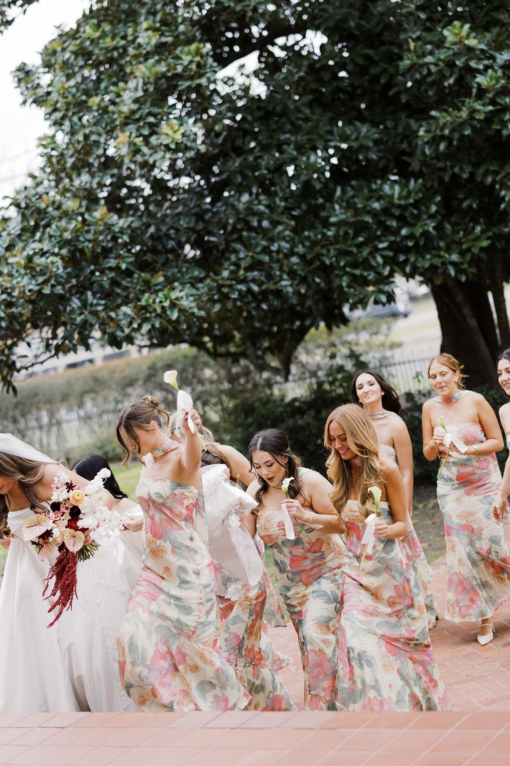 Bridesmaids walking together while holding white calla lilies and helping the bride with her dress.