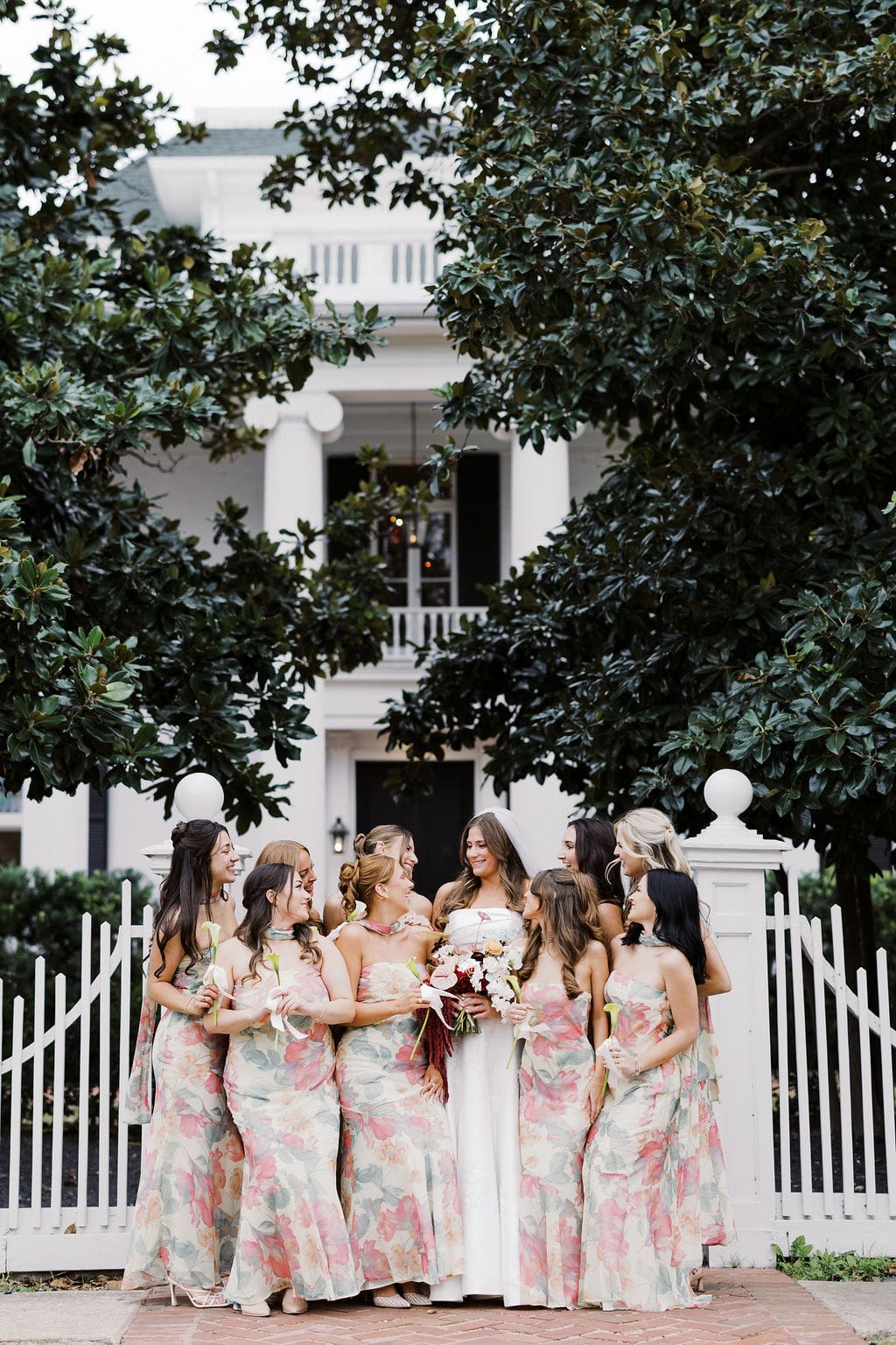 Bride and bridesmaids in floral dresses gathered in front of a white estate entrance during a joyful Mansion Wedding portrait.