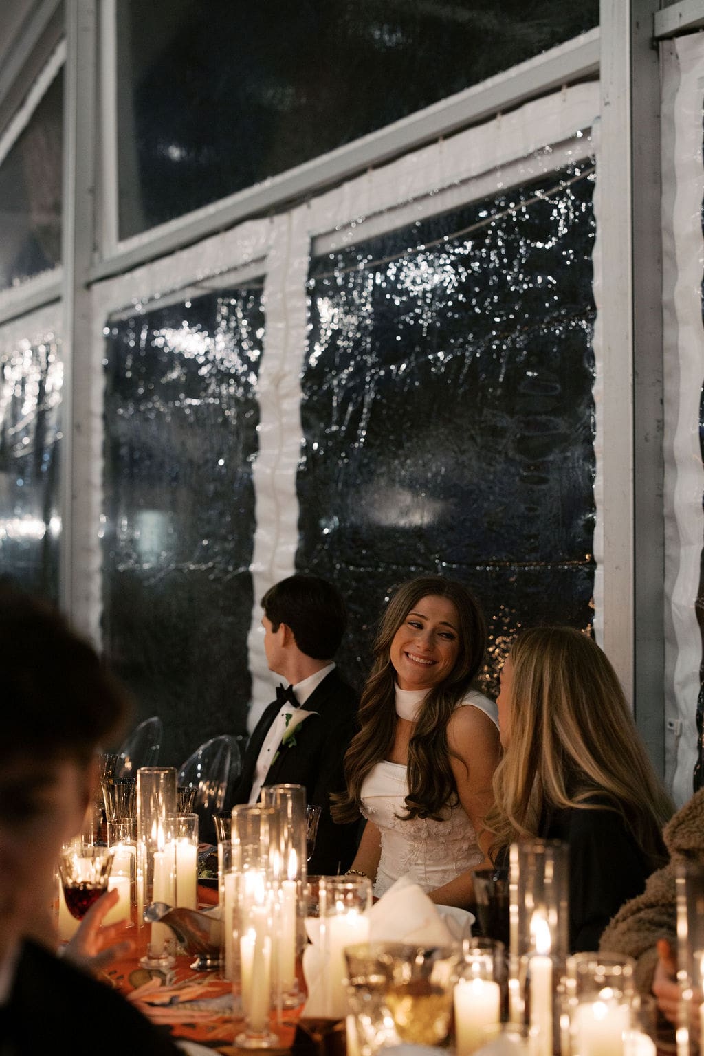 Bride smiling while seated at a candlelit reception table surrounded by guests and glowing glass candle holders.