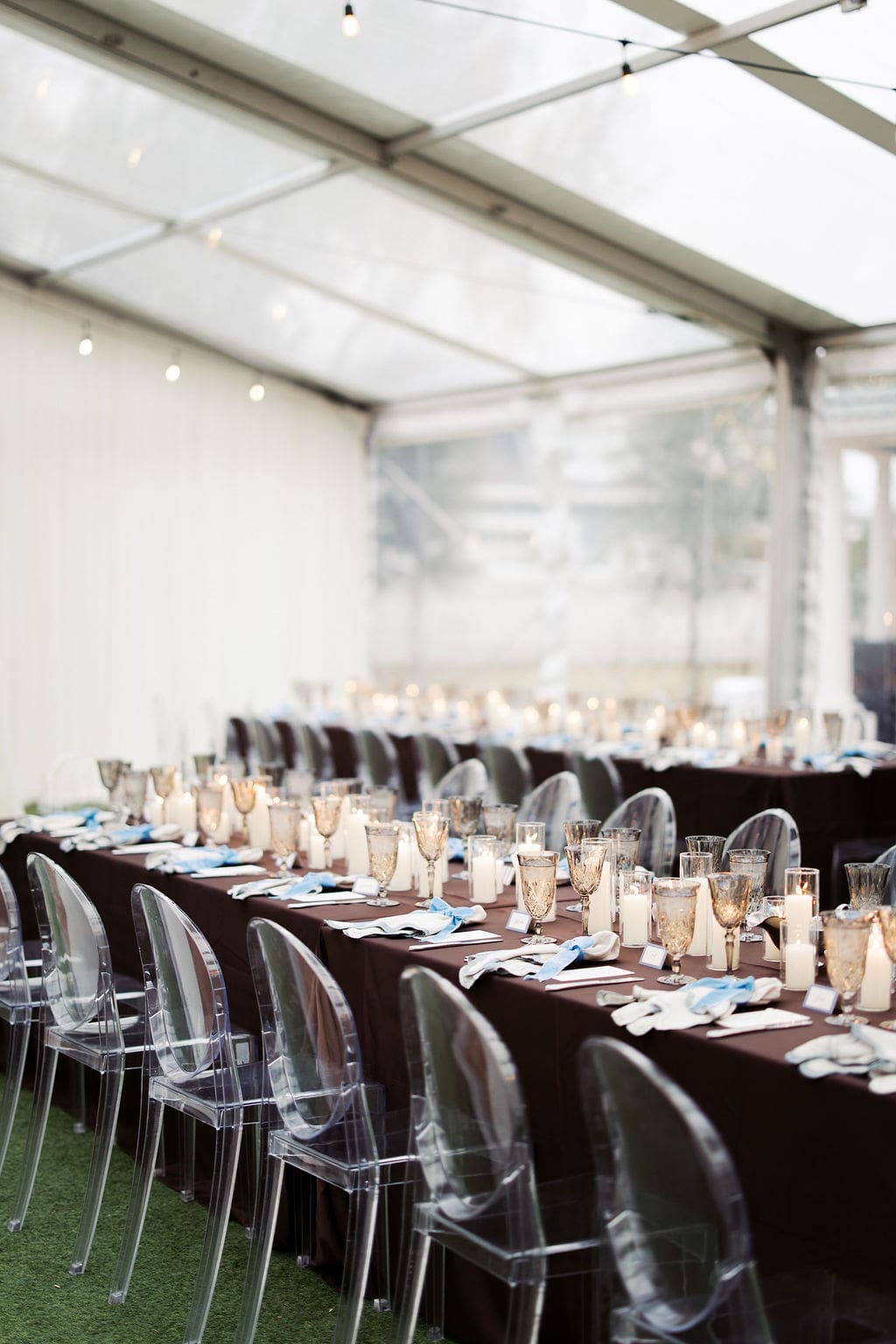 Elegant reception tables with brown linens, clear chairs, and candlelight inside a tent during a stylish Mansion Wedding reception.