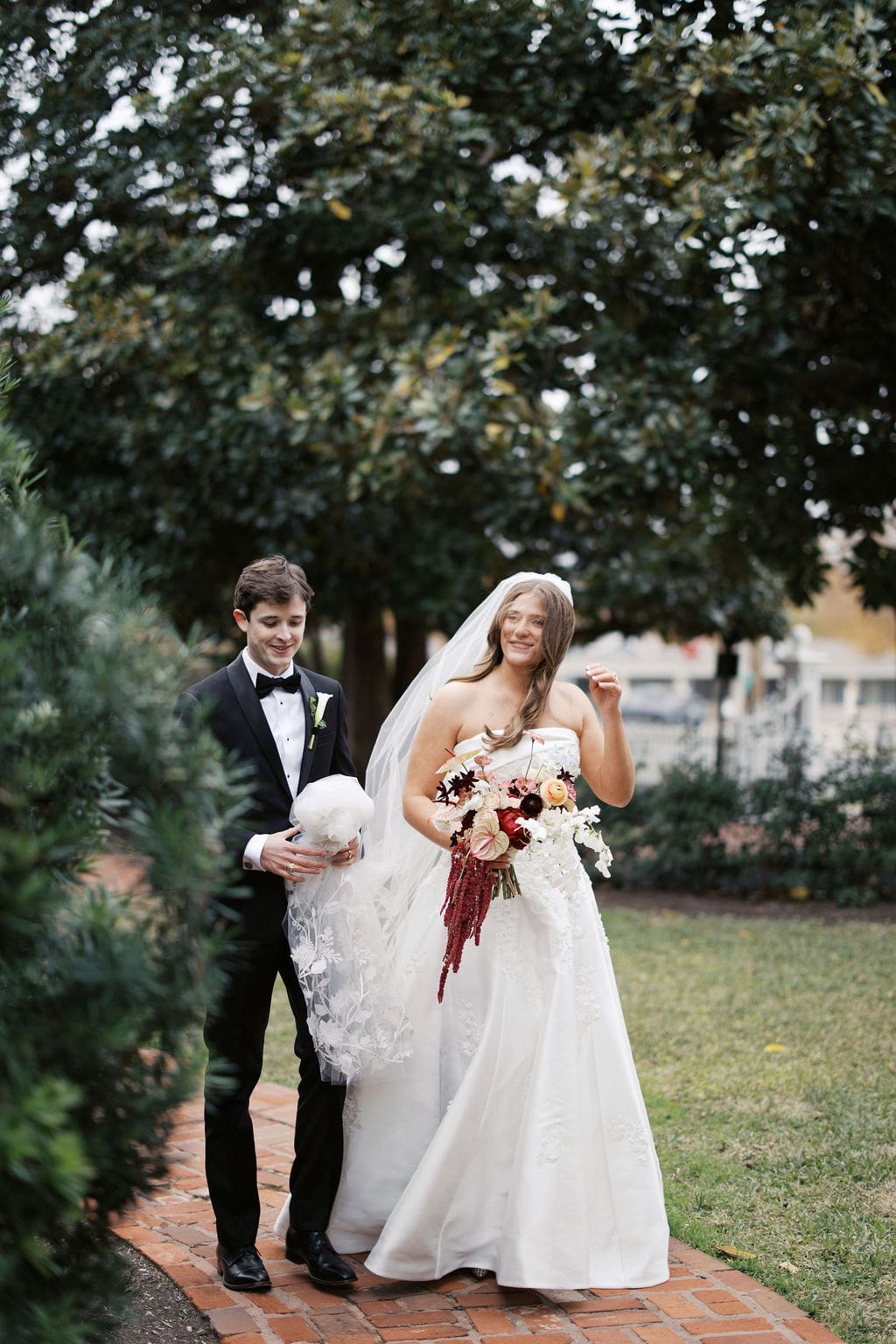 Bride and groom walking together along a garden path while holding a dramatic red and blush bouquet.