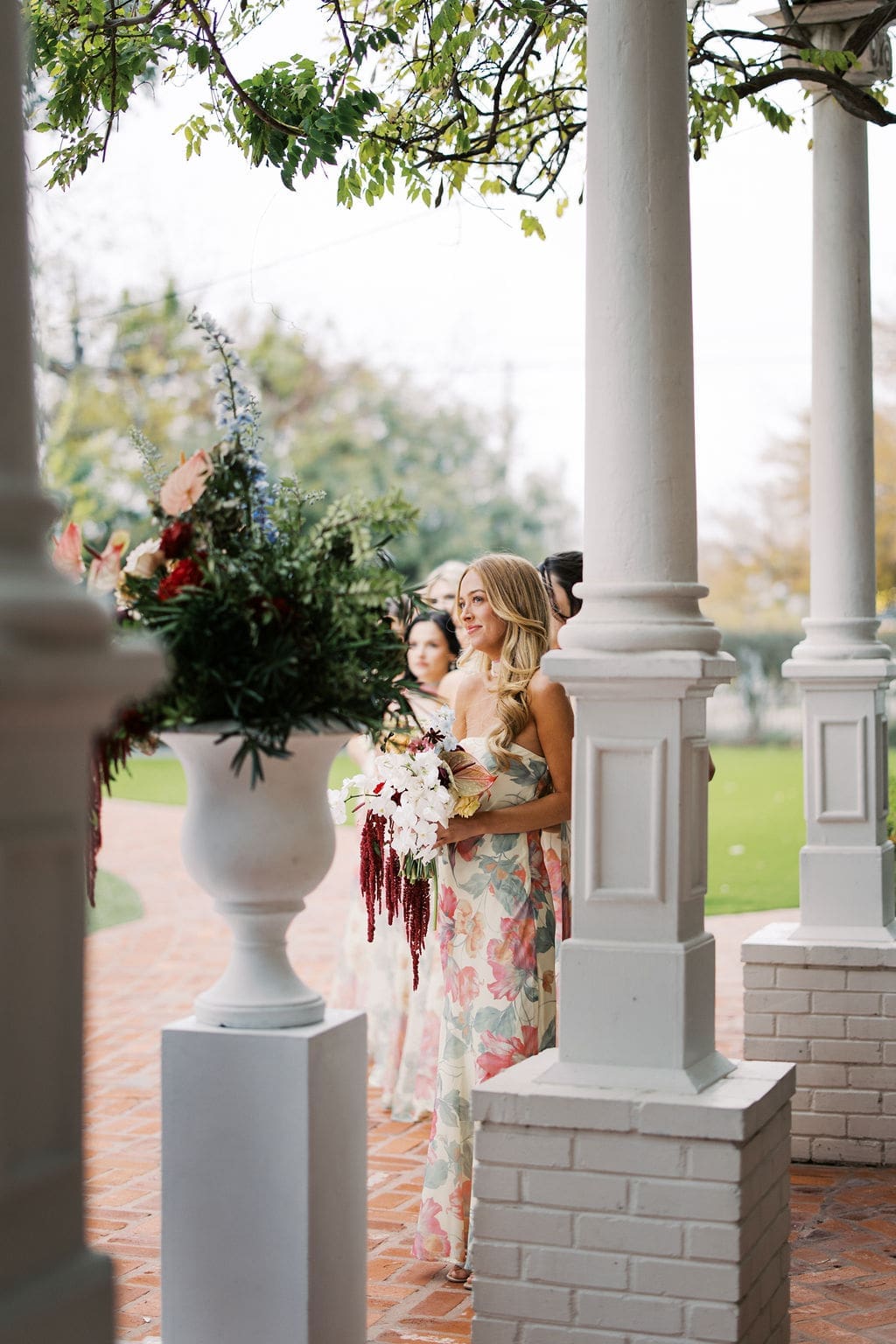 Bridesmaid holding a bouquet while waiting beneath white porch columns before the ceremony begins.