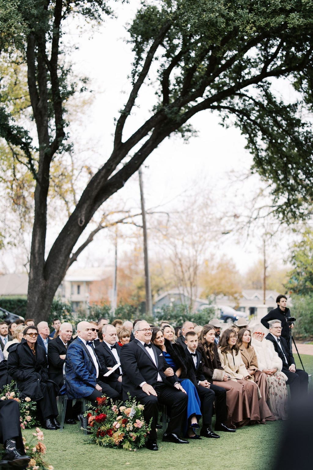 Guests seated outdoors beneath large trees during a heartfelt Mansion Wedding ceremony.