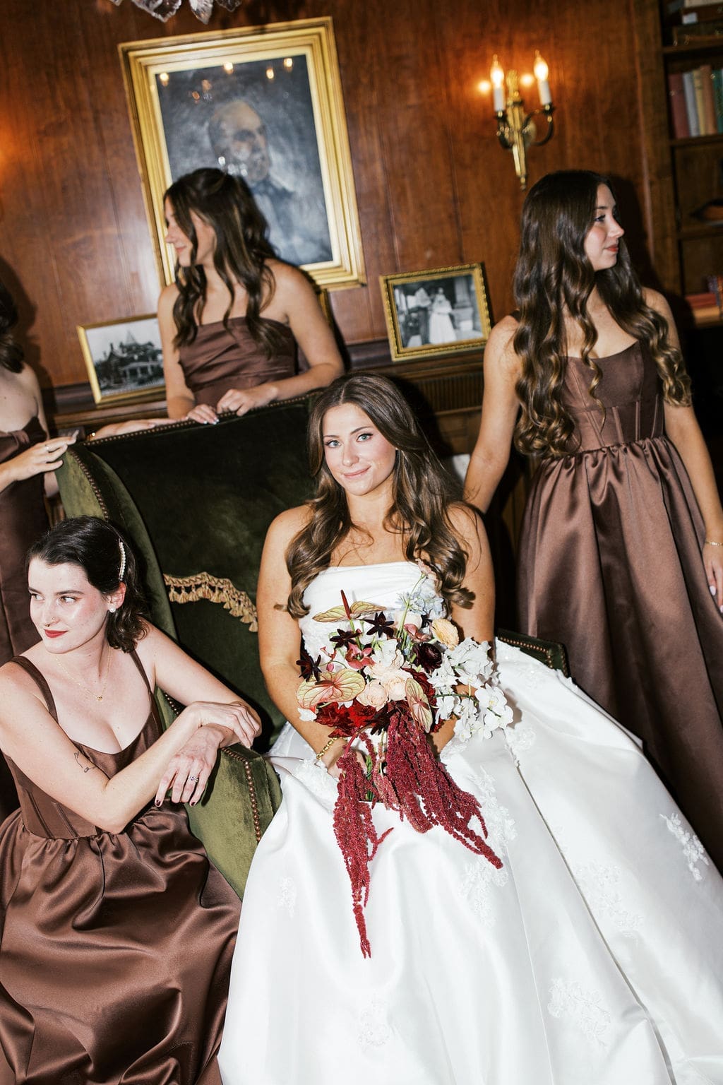 Bride sitting in a velvet chair surrounded by bridesmaids in chocolate satin dresses during relaxed bridal party portraits indoors.