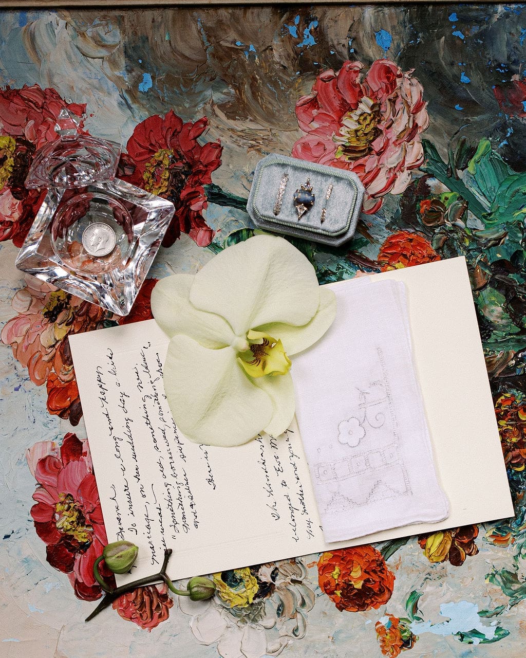 Flat lay of heirloom wedding details including rings, perfume bottle, handwritten note, and floral accents on a colorful painted background.