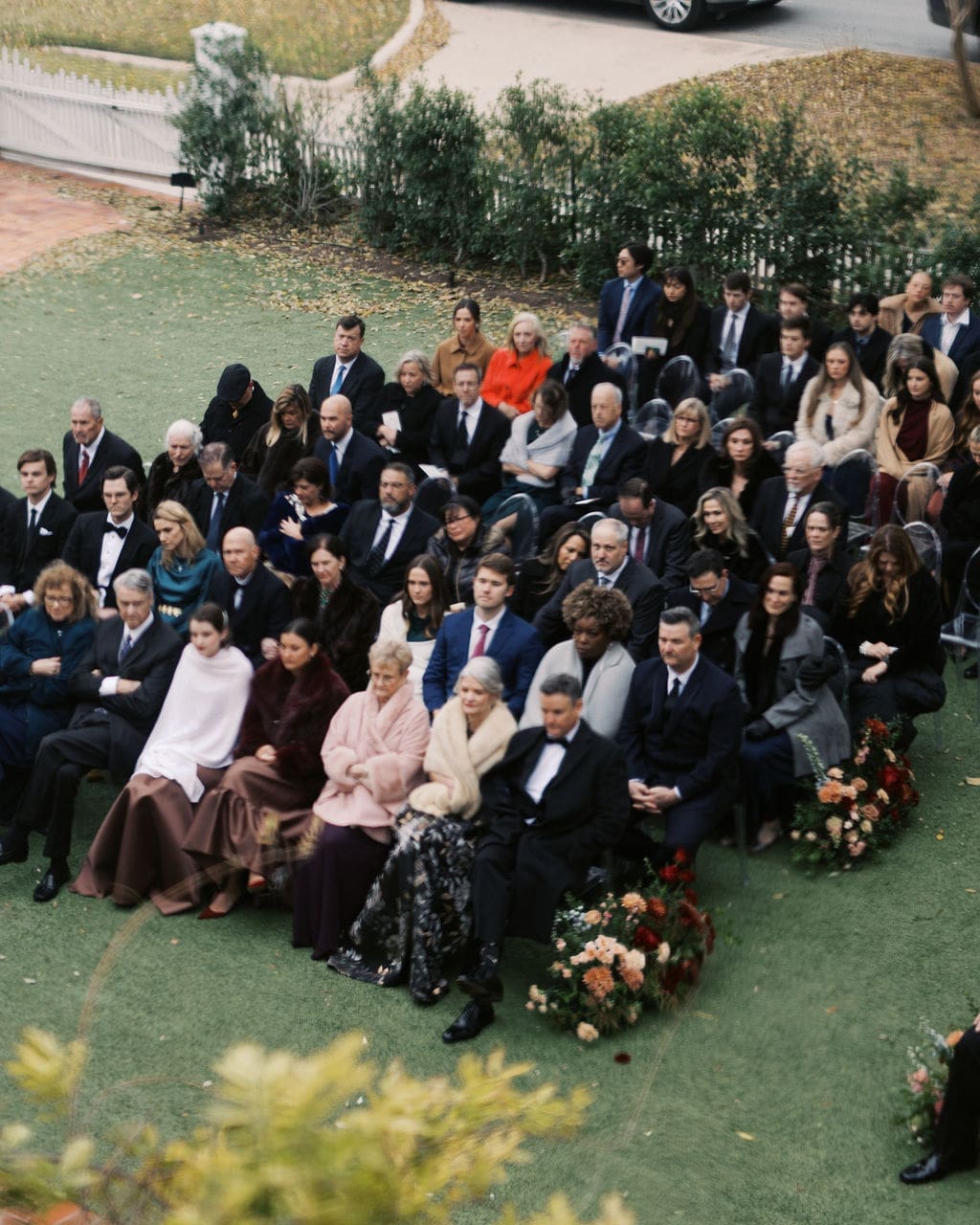 Guests seated in clear ceremony chairs outdoors, waiting for the wedding ceremony to begin in a garden setting.