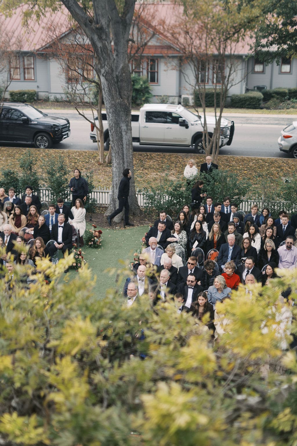 Wedding guests seated outdoors watching the ceremony from clear chairs beneath autumn trees.