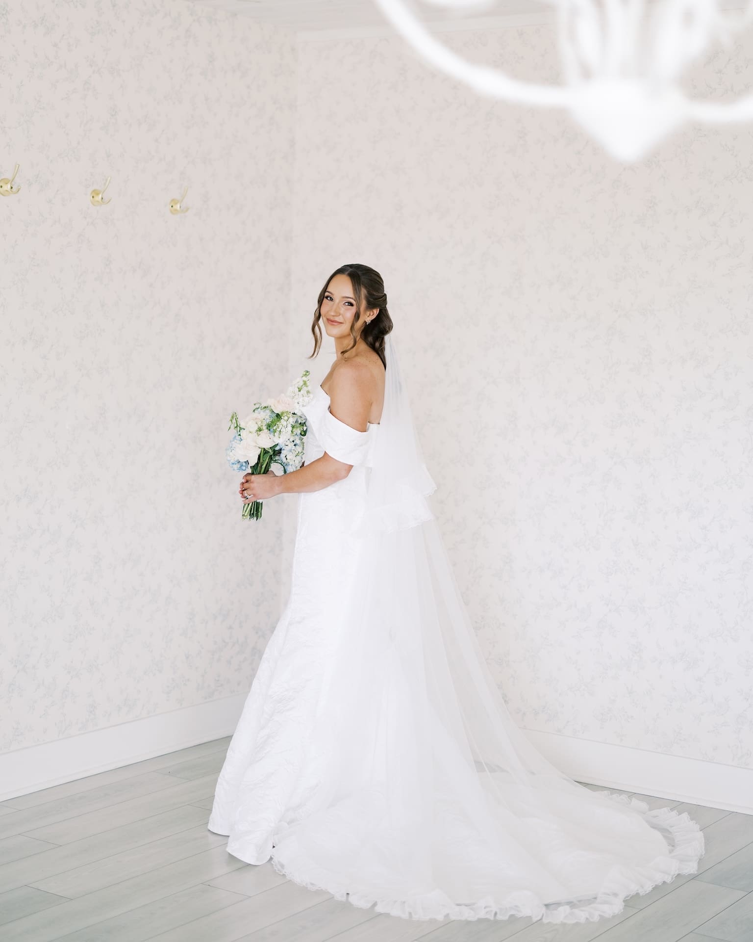 Elegant bridal portraits of a bride in an off-the-shoulder gown with a cathedral veil and tiered ruffled train, smiling back at the camera in a softly lit floral wallpapered room