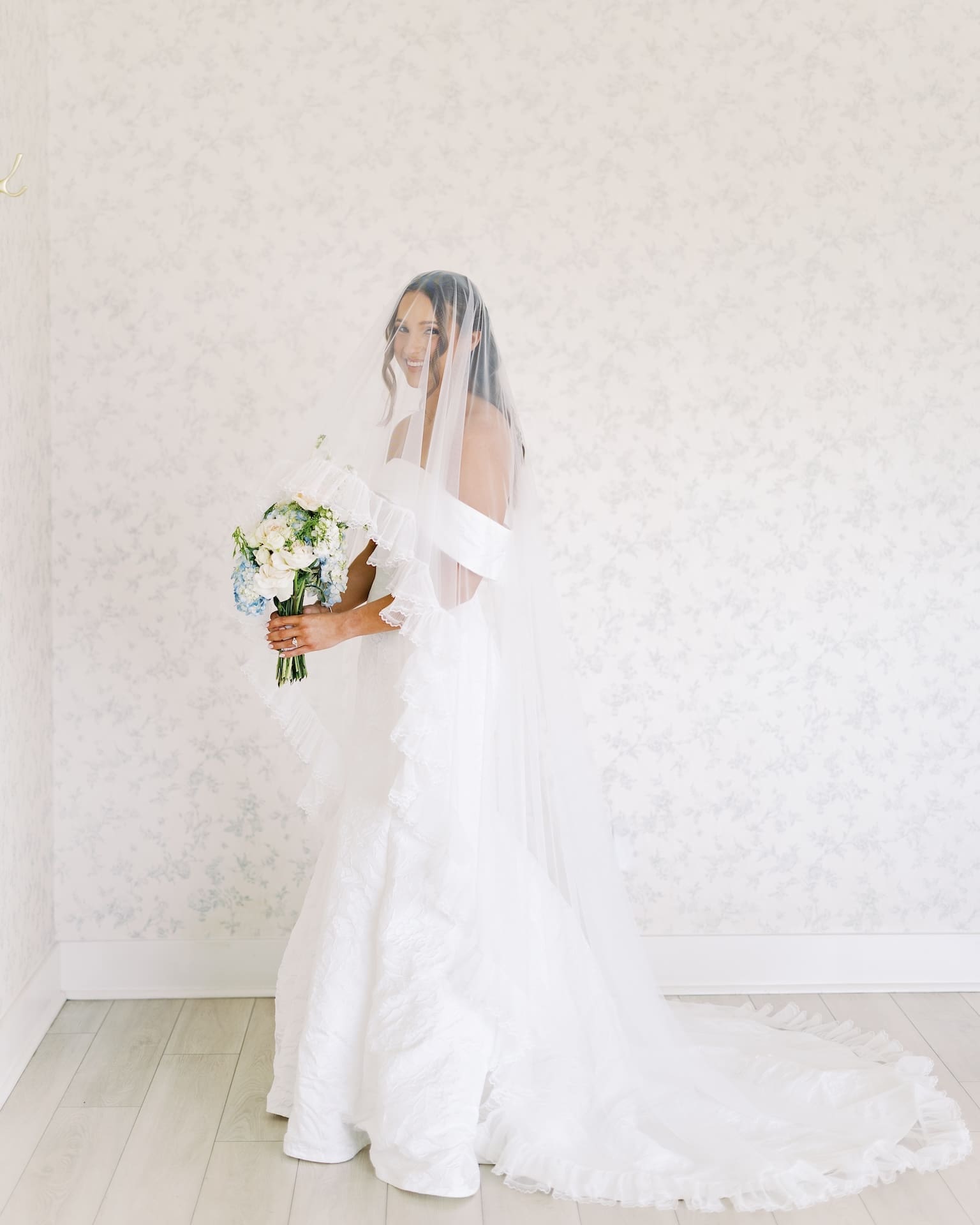 Dreamy bridal portraits of a bride in an off-the-shoulder gown and ruffled veil draped over her face, smiling as she stands in profile against soft floral wallpaper holding a blue and white bouquet