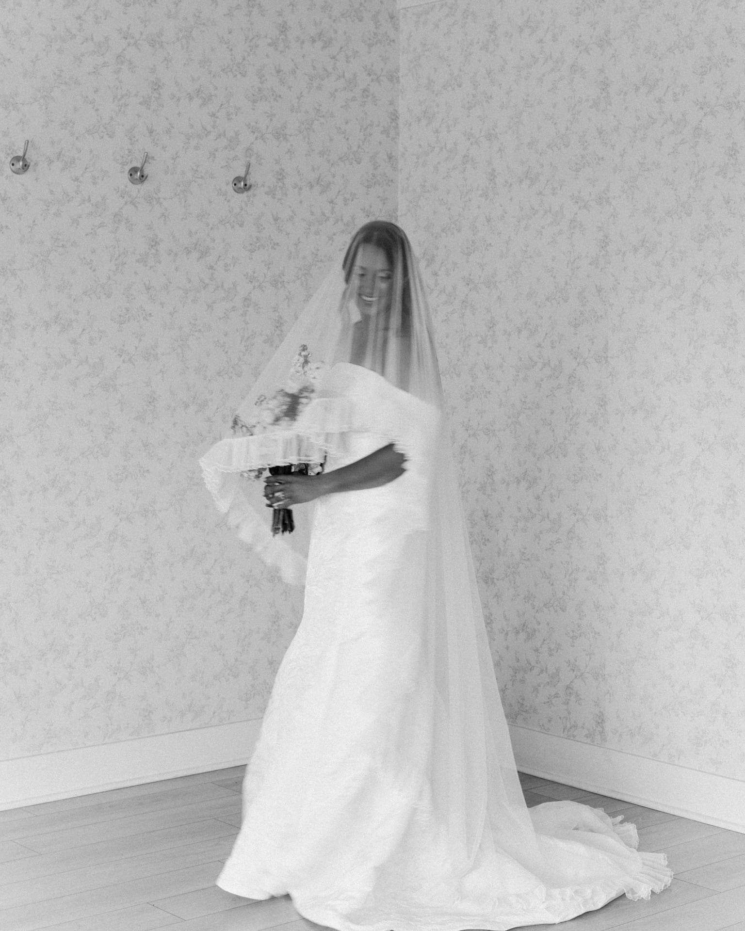 Moody black and white bridal portraits of a bride standing in the corner of a floral wallpapered room, veil draped over her face, motion blur adding a dreamy effect