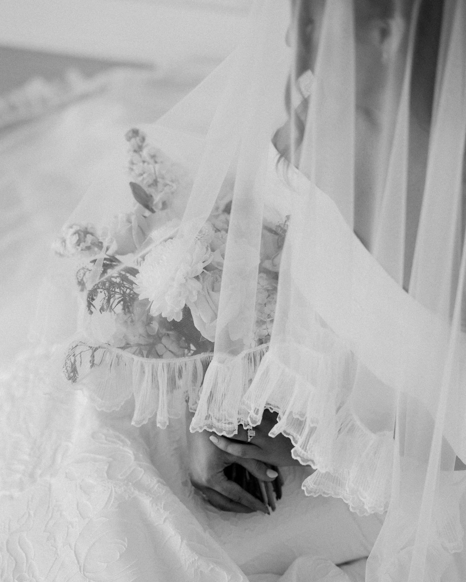 Black and white close-up of a bride's hands resting over her bouquet, the ruffled lace edge of her veil cascading over the flowers and her lace gown in a dreamy detail shot