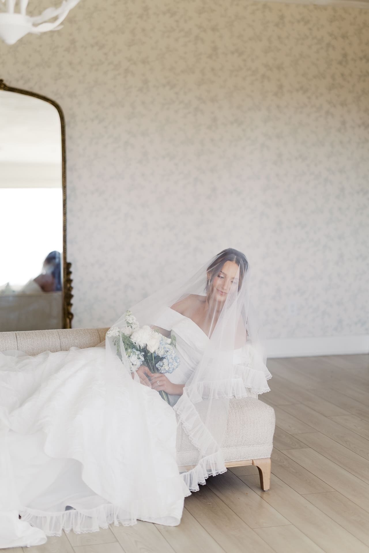 A bride sits on a settee in a light-filled bridal suite, veil cascading over her as she gazes down at her bouquet, a gilded mirror reflecting the room behind her