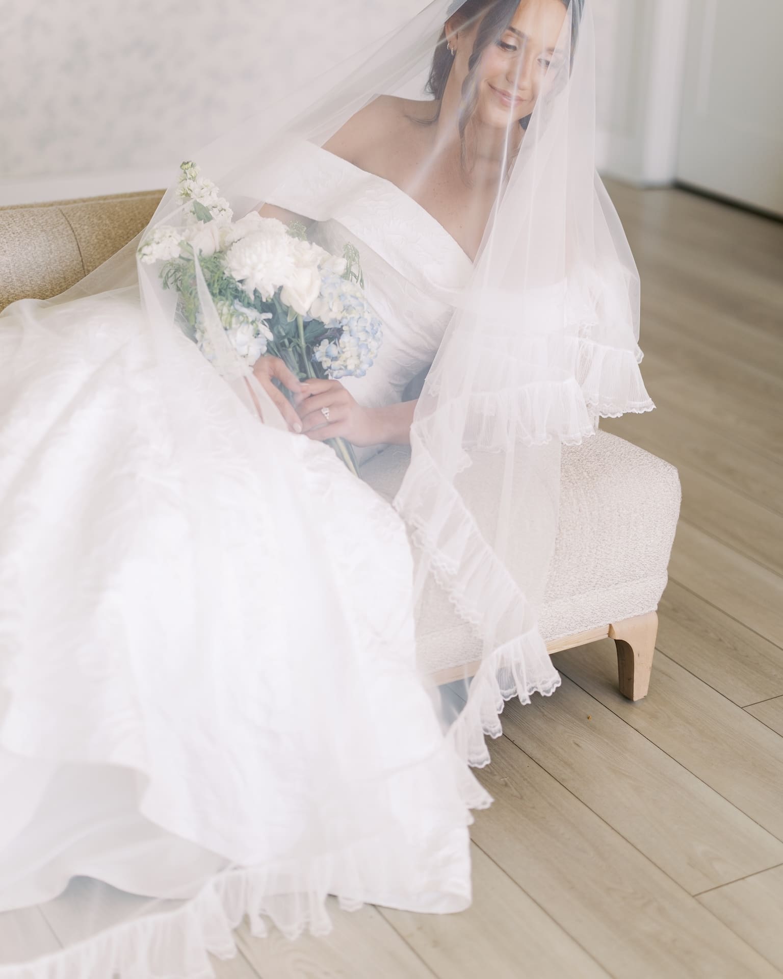 A bride sits on a cream settee beneath her flowing veil, holding a bouquet of white peonies and blue hydrangeas, her engagement ring catching the light