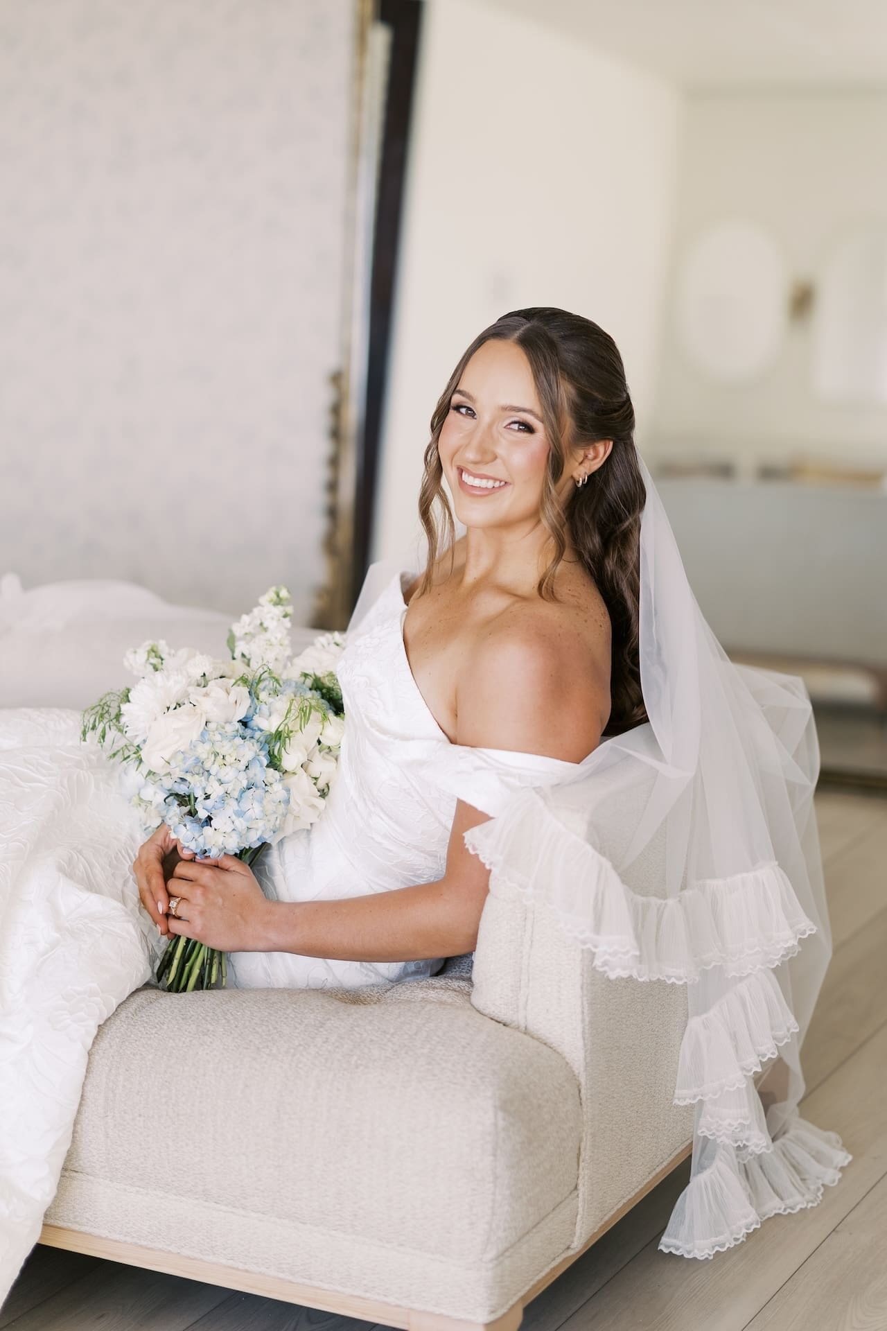 A bride sits on a cream settee in a bright bridal suite, laughing and holding a lush bouquet of blue hydrangeas and white blooms, a gilded mirror reflecting the room behind her