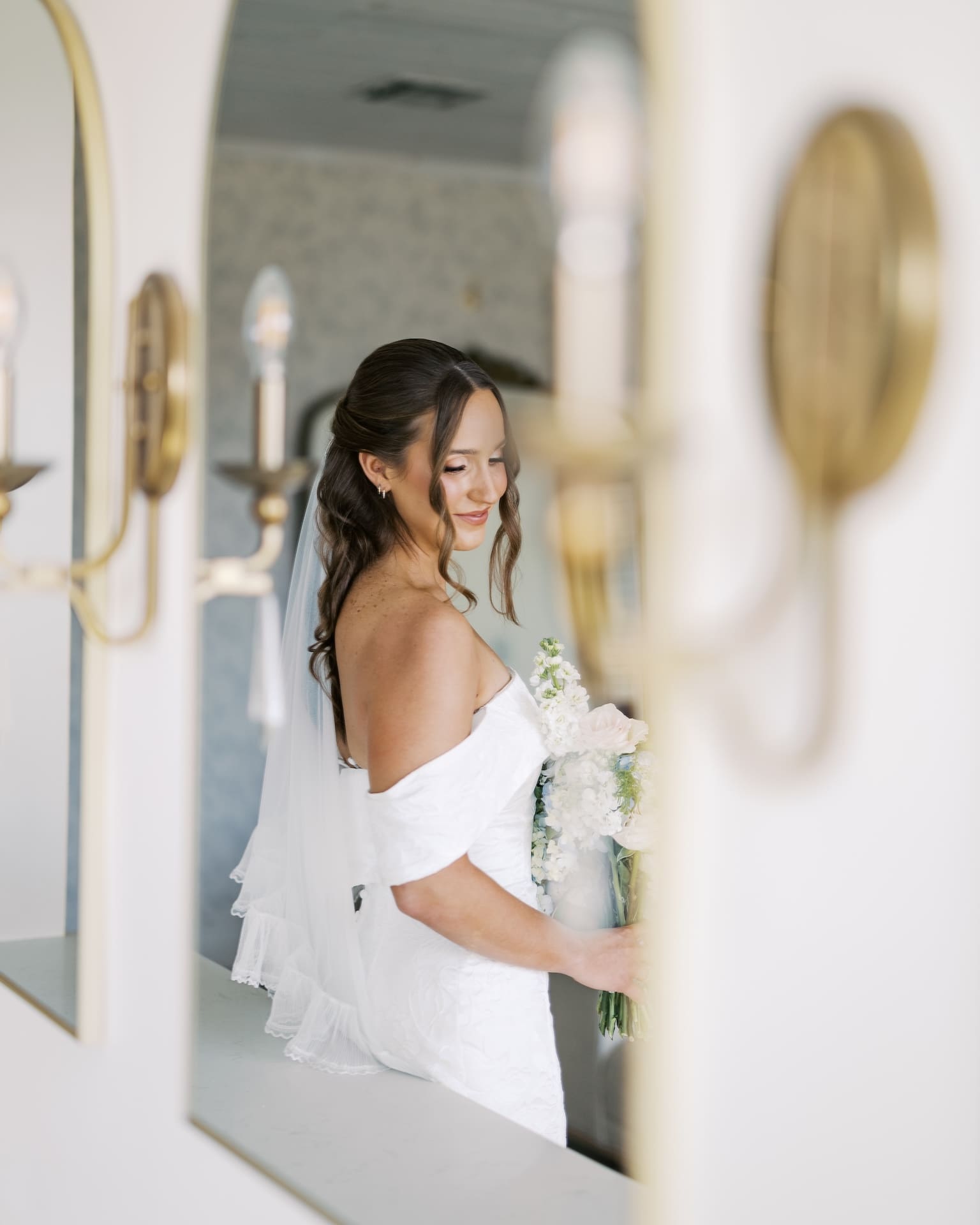 Artistic bridal portraits shot through a doorway framed by brass sconces, capturing a bride in an off-the-shoulder gown gazing down at her bouquet in soft natural light