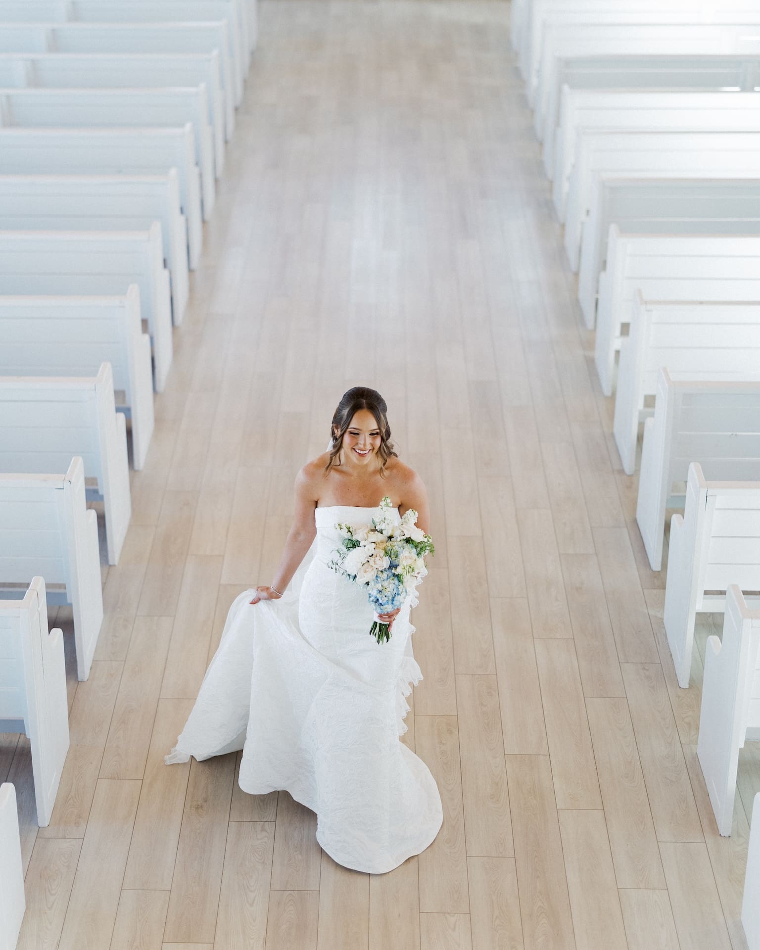 Aerial view looking down the chapel aisle as a bride in a strapless gown walks forward smiling, her ruffled train fanning out across the light hardwood floor