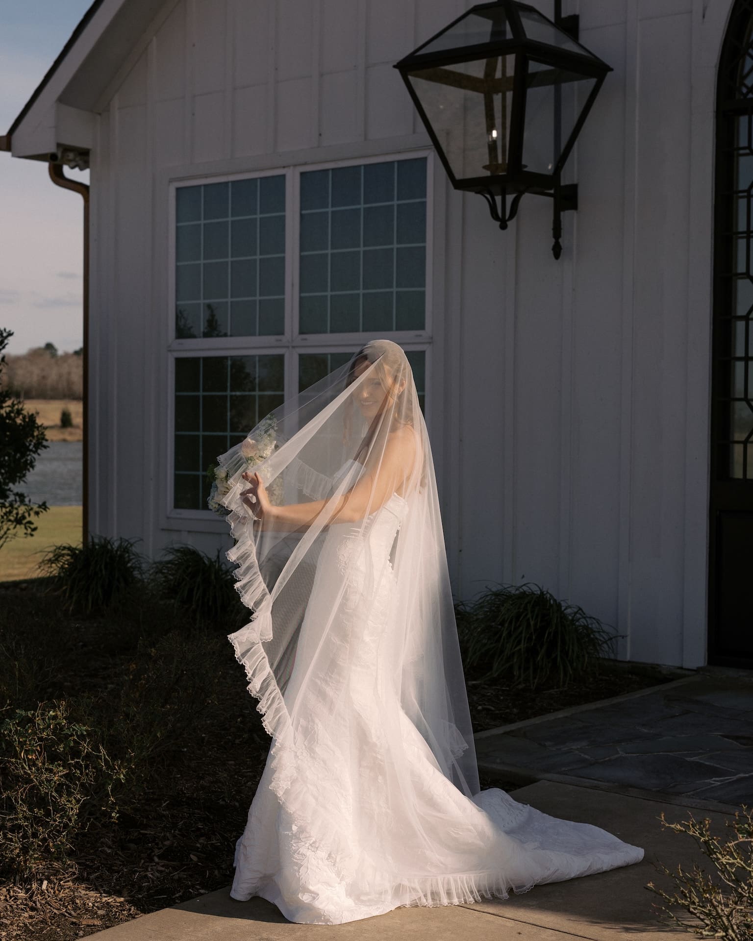 A bride stands outside a white barn chapel at golden hour, her long ruffled veil draped over her face as she holds a small bouquet