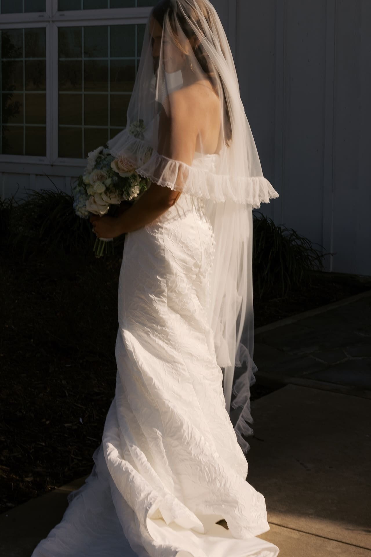 A bride in an off-the-shoulder gown stands in dramatic side lighting outside a white barn venue, her ruffled veil cascading down her back as she holds a garden bouquet
