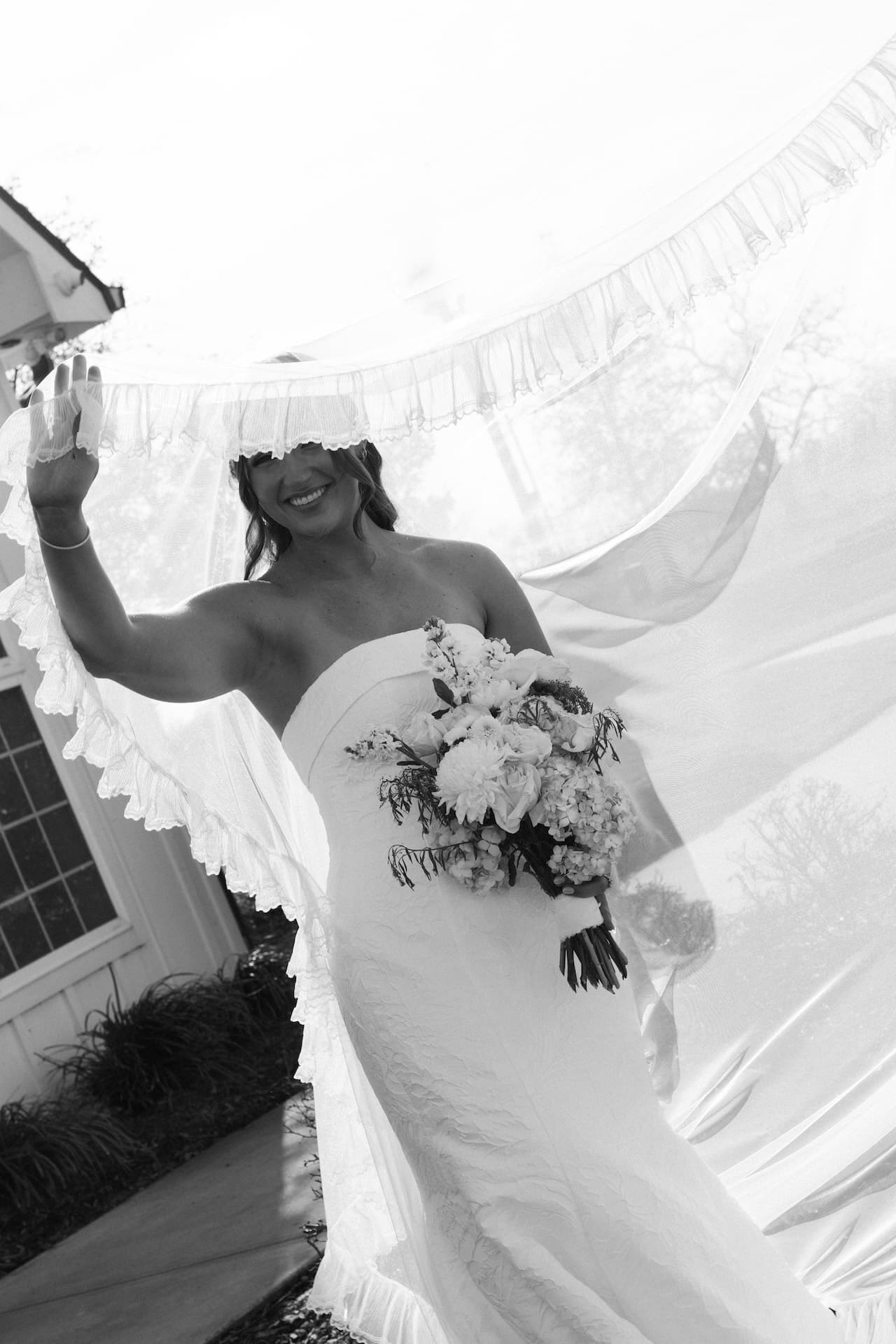 Black and white photo of a bride laughing joyfully as her ruffled veil billows around her in the wind, bouquet in hand outside a white chapel